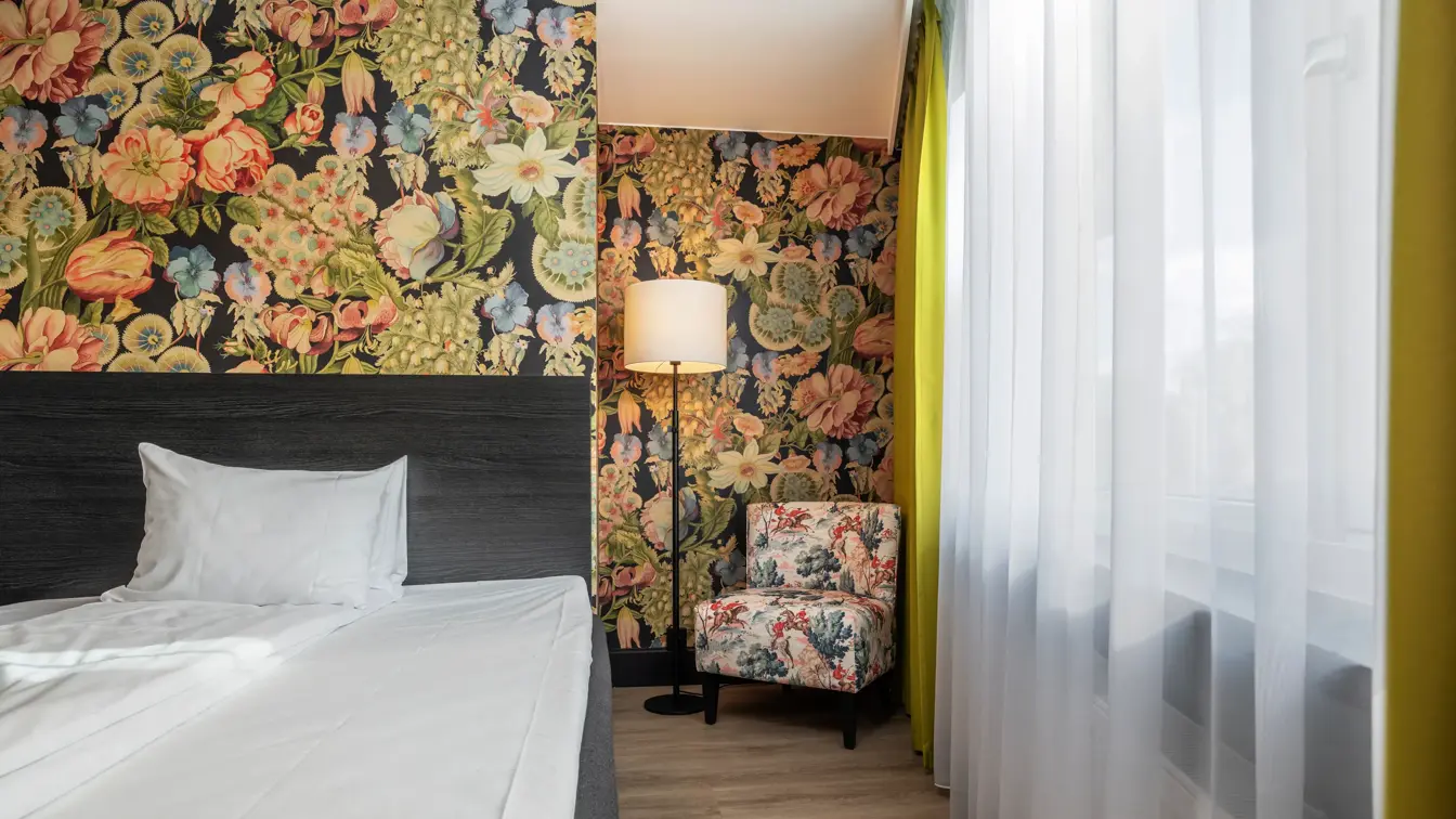 Enkeltrom med enkeltseng, fargerik tapet, stort vindu, lenestol og stålampe.