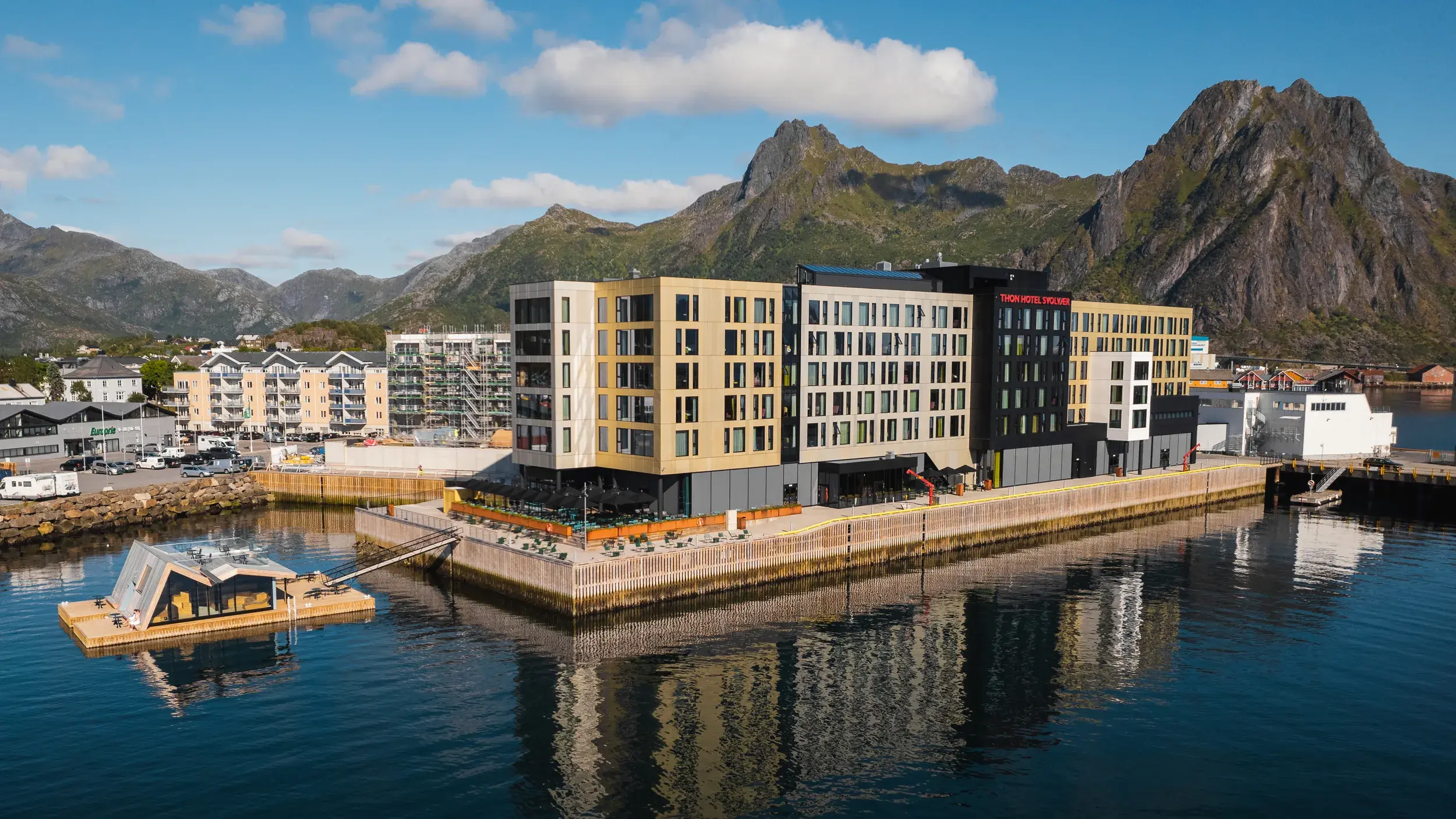 Moderne bygning på kaiområde som speiler seg i vannflaten, med utendørs sauna nedenfor og grønne fjell i bakgrunnen.