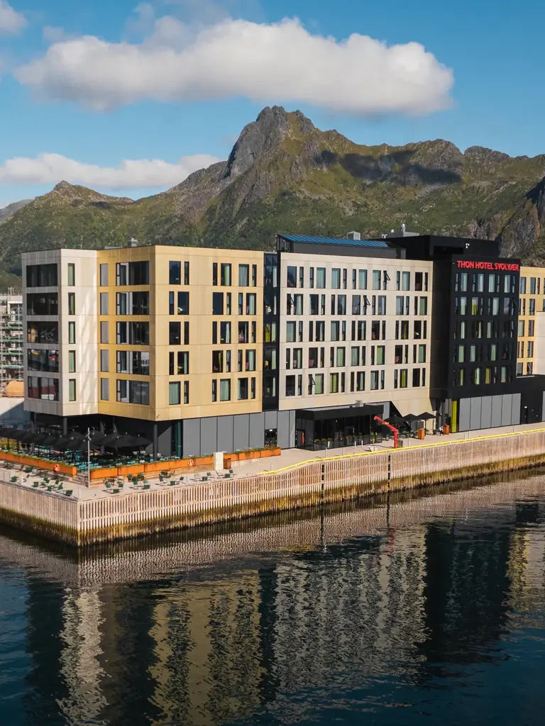 Moderne bygning på kaiområde som speiler seg i vannflaten, med utendørs sauna nedenfor og grønne fjell i bakgrunnen.