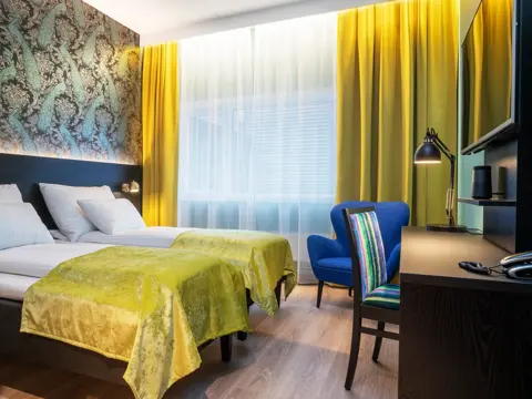 Gule gardiner, vindu fra gulv til tak, to enkel senger, tapetvegg me dmønster, skrivebord, TV, blå stol, skrivebordstol, parkettgulv. 