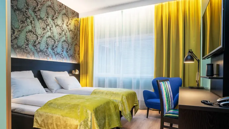 Gule gardiner, vindu fra gulv til tak, to enkel senger, tapetvegg me dmønster, skrivebord, TV, blå stol, skrivebordstol, parkettgulv. 