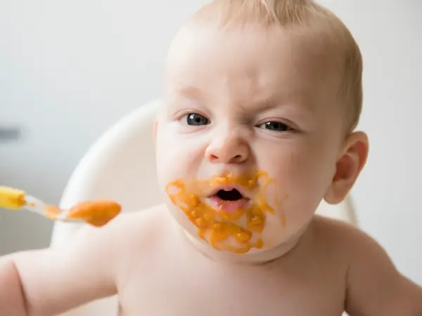 Mother feeding messy baby son with spoon in high chair