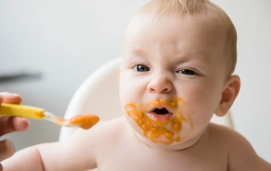 Mother feeding messy baby son with spoon in high chair