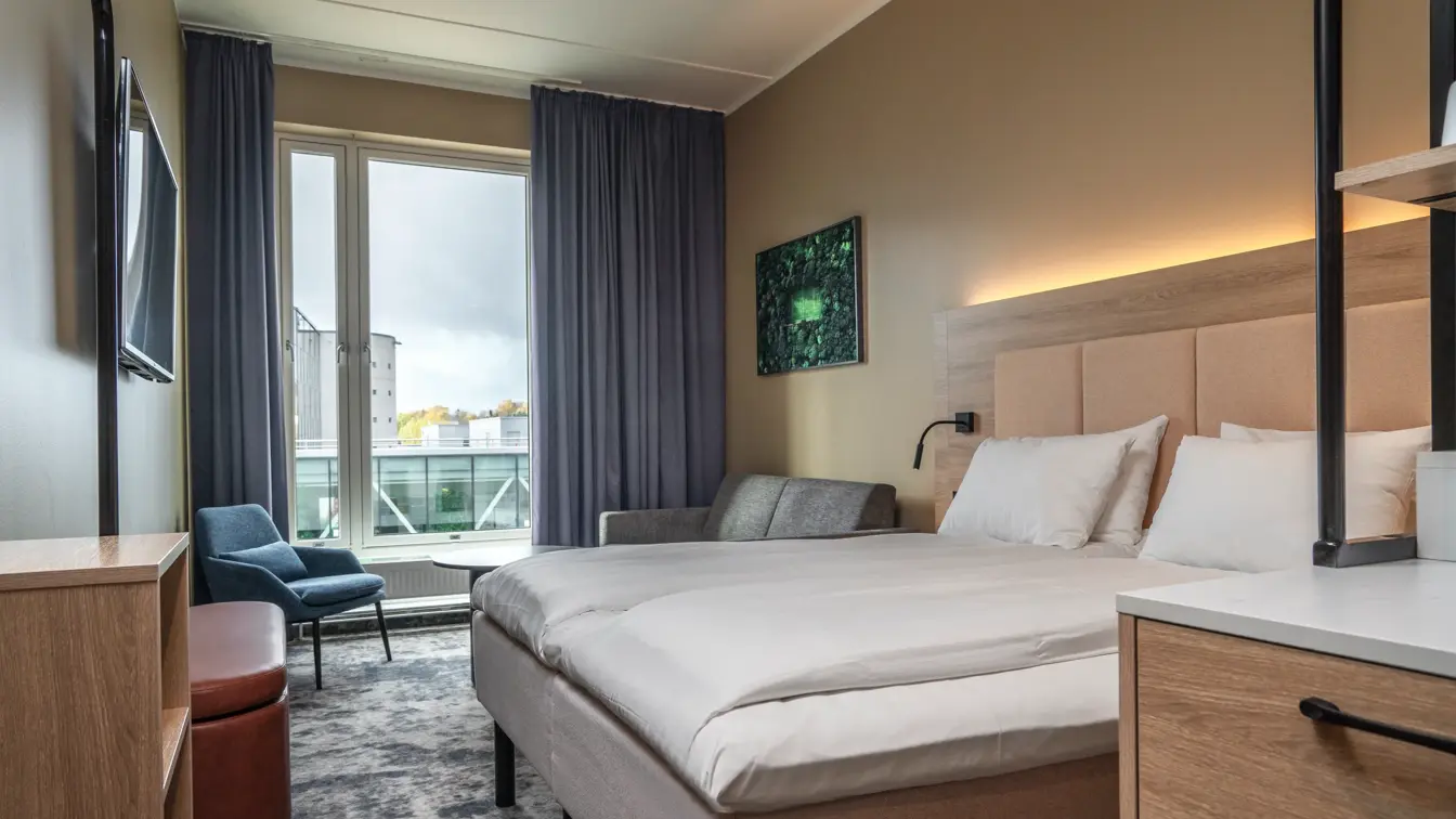 Standard dobbeltrom med dobbeltseng, sittegruppe, vegghengt TV, stort vindu med gardiner på hver side.