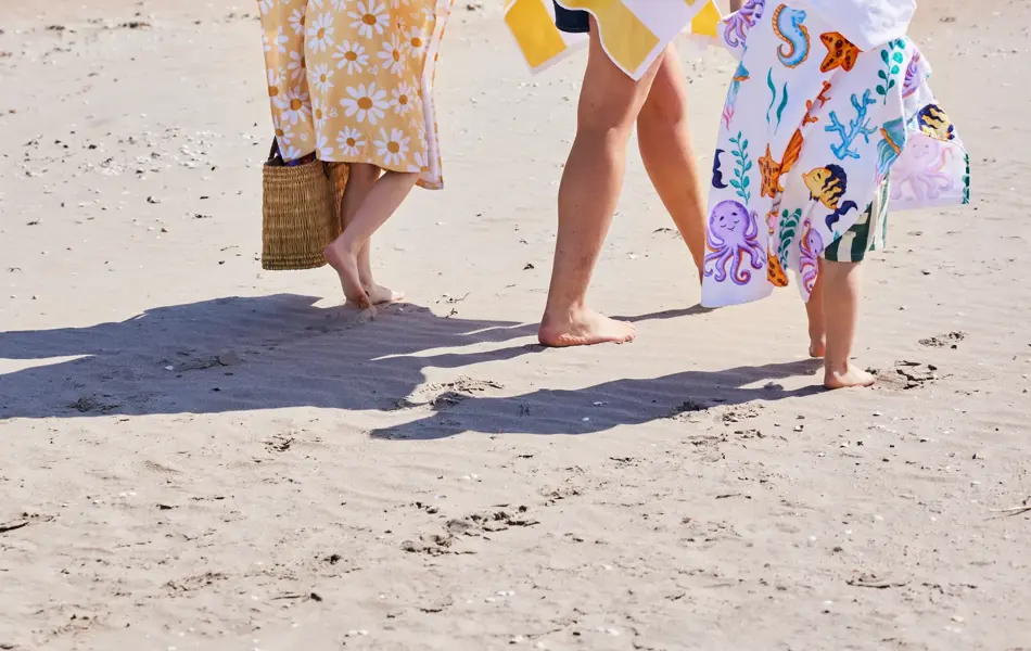 Dame med to barn som går på stranden med håndklær rundt seg