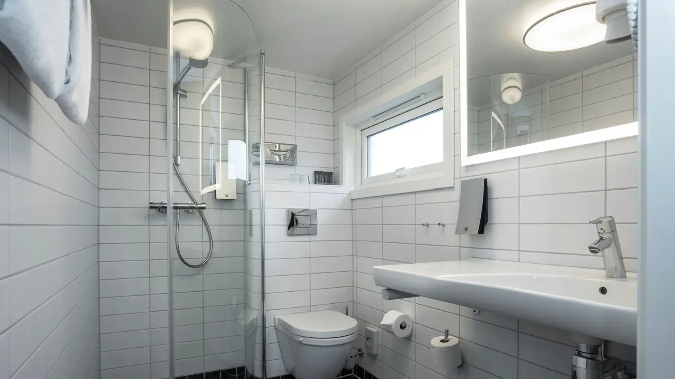 Golv med små sorte fliser og vegger med større hvite fliser. Vegghent servant og speil med led lys rundt. Lite vindu, vegghent toalett og glassvegg til dusjhjørne. 