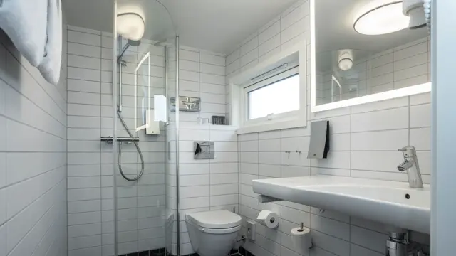 Golv med små sorte fliser og vegger med større hvite fliser. Vegghent servant og speil med led lys rundt. Lite vindu, vegghent toalett og glassvegg til dusjhjørne. 
