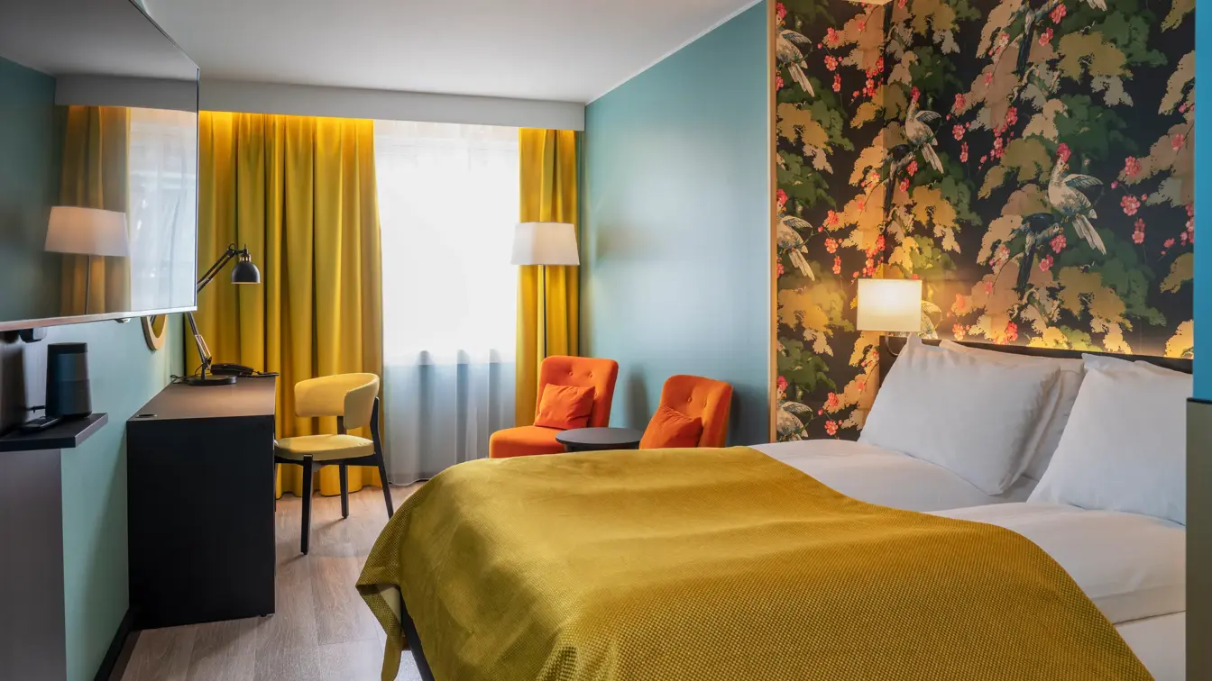 Fargerik tapet og dobbeltseng med gult sengeteppe. Turkis tapet, oransje stoler, mørkt rundt bord og gule gardiner. Skrivebord med leselampe og gul stol i dobbeltrom på Thon Hotel Bristol Bergen