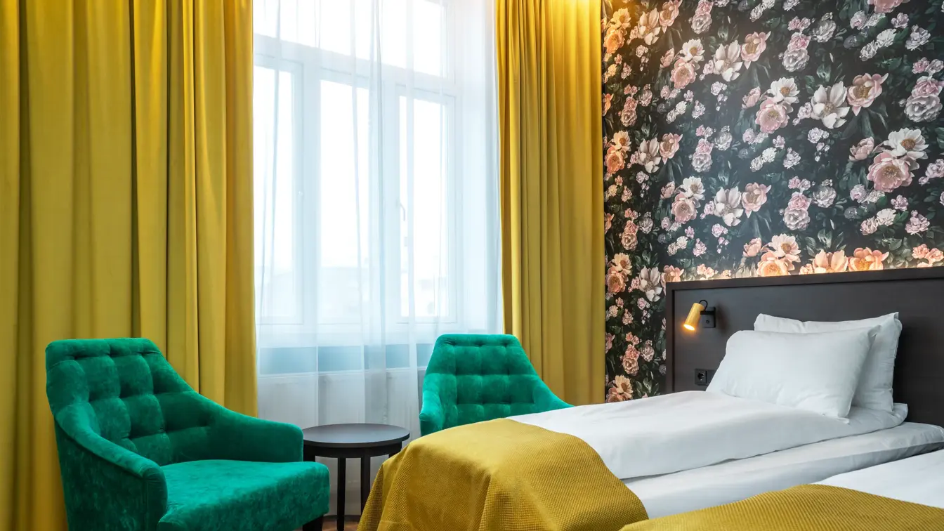Stort vindu med gule gardiner fra tak til gulv. To grønne lenestoler og et lite rundt mørkt bord. Mørk tapet med store lyserosa roser. To enkeltsenger med gule sengetepper. I standard twin rom på Thon Hotel Høyers i Skien.