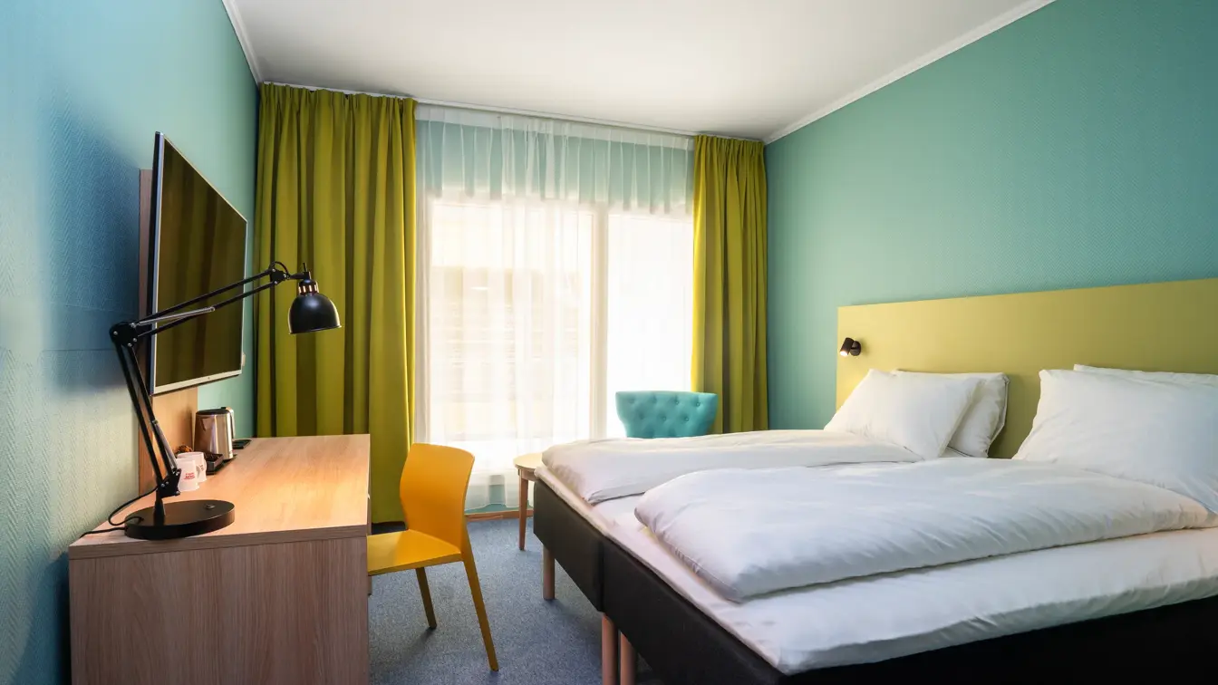 Skrivebord, stol, TV, gule gardiner, veggspeil, dobbeltseng, turkise vegger, turkis stol. 