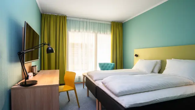 Skrivebord, stol, TV, gule gardiner, veggspeil, dobbeltseng, turkise vegger, turkis stol. 
