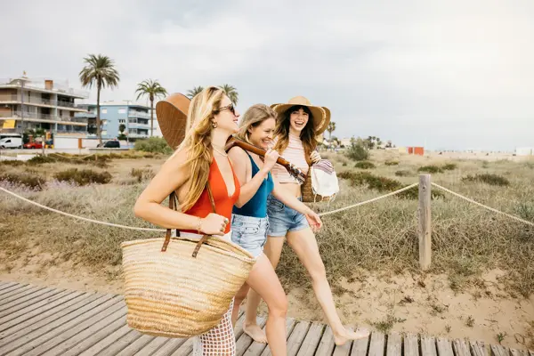 Tre damer med solhatt, gitar og stor stråveske går på på stranden sett fra siden 