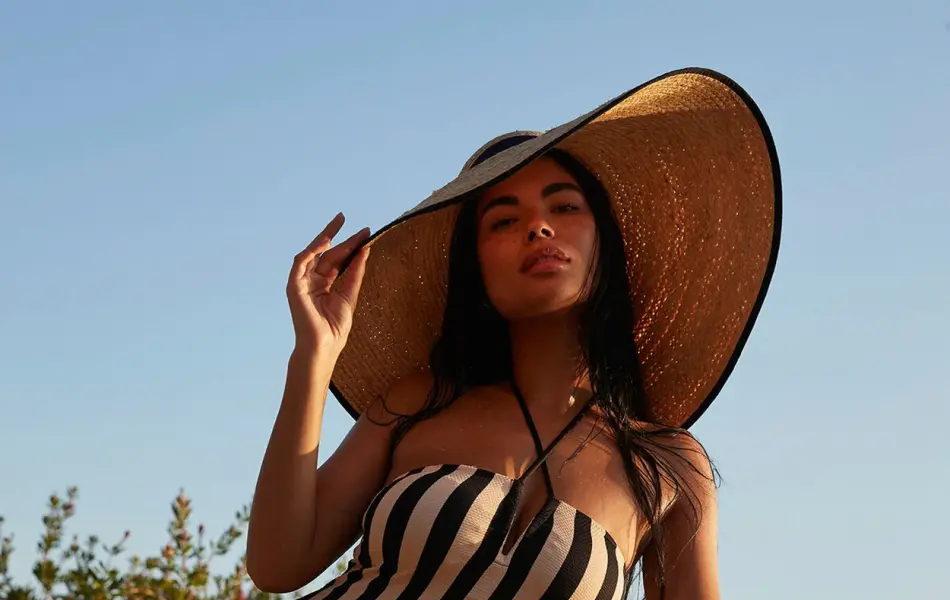 Modell står ved ute ved en badetrapp med stor solhatt og stripete badedrakt.