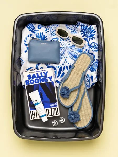 Boks med blåmønstret kjole, hvite solbriller, blå lommebok, Sally rooney bok, kremtube, øredobber og filp flops.