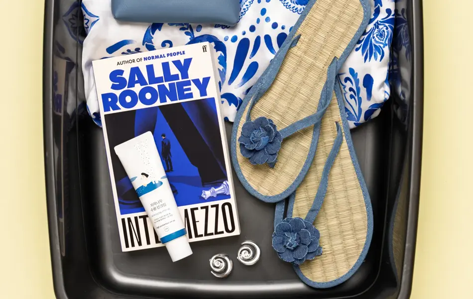 Boks med blåmønstret kjole, hvite solbriller, blå lommebok, Sally rooney bok, kremtube, øredobber og filp flops.