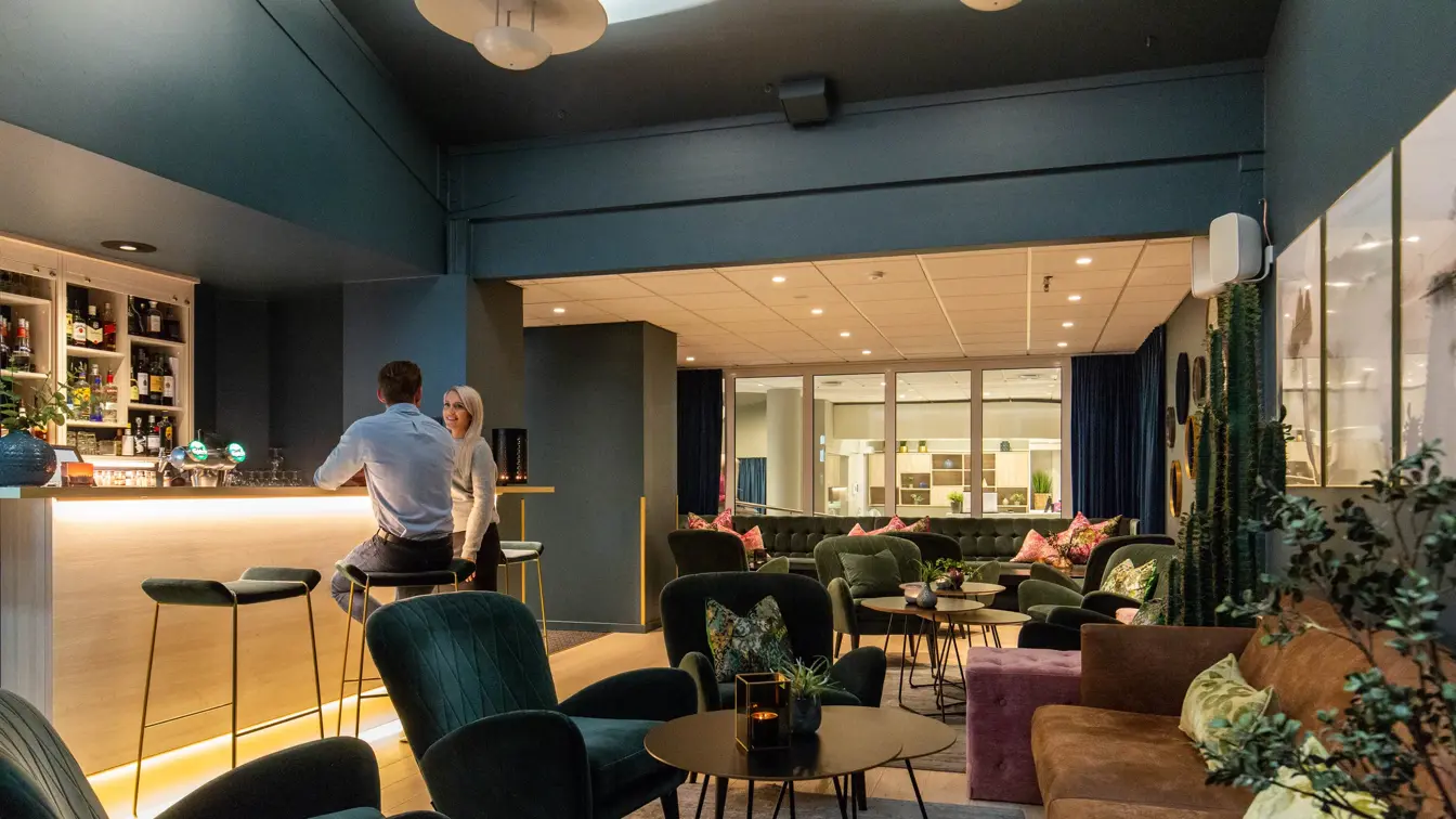 Bar, grønne stoler, plantter, sofa, pynteputer, bord, to menn i baren