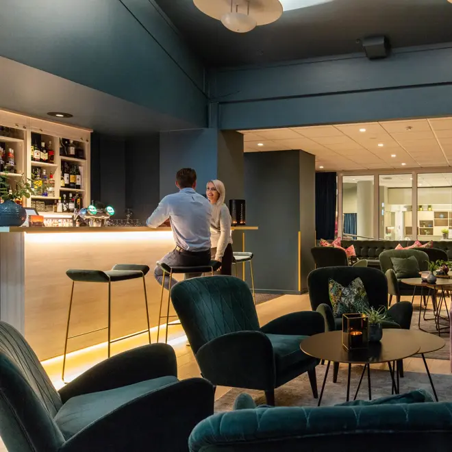 Bar, grønne stoler, plantter, sofa, pynteputer, bord, to menn i baren