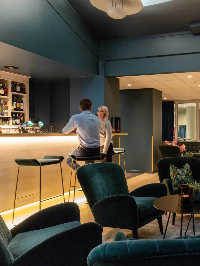 Bar, grønne stoler, plantter, sofa, pynteputer, bord, to menn i baren