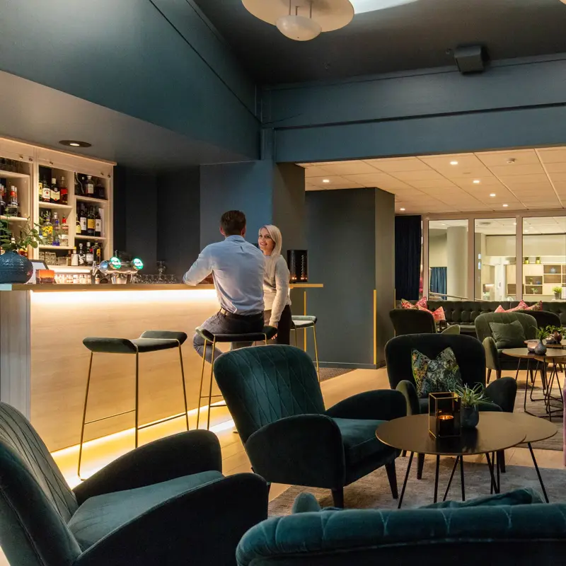 Bar, grønne stoler, plantter, sofa, pynteputer, bord, to menn i baren