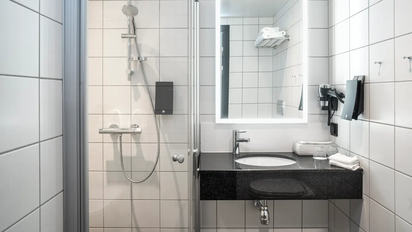 Hvite veggfliser, svarte gulvfliser, dusj, vask, speil, såpe.