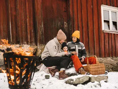 To voksne og et barn sitter ute og smiler inntil en rød vegg i snøen, ved et bål