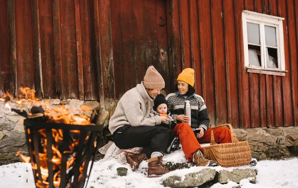 To voksne og et barn sitter ute og smiler inntil en rød vegg i snøen, ved et bål