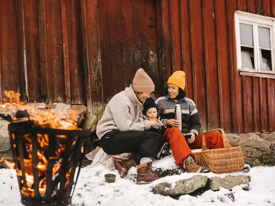 To voksne og et barn sitter ute og smiler inntil en rød vegg i snøen, ved et bål