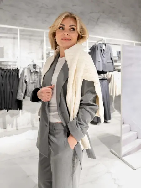 Dame i grå dressbukse og jakke, hvit topp og hvit strikkegenser over skuldre