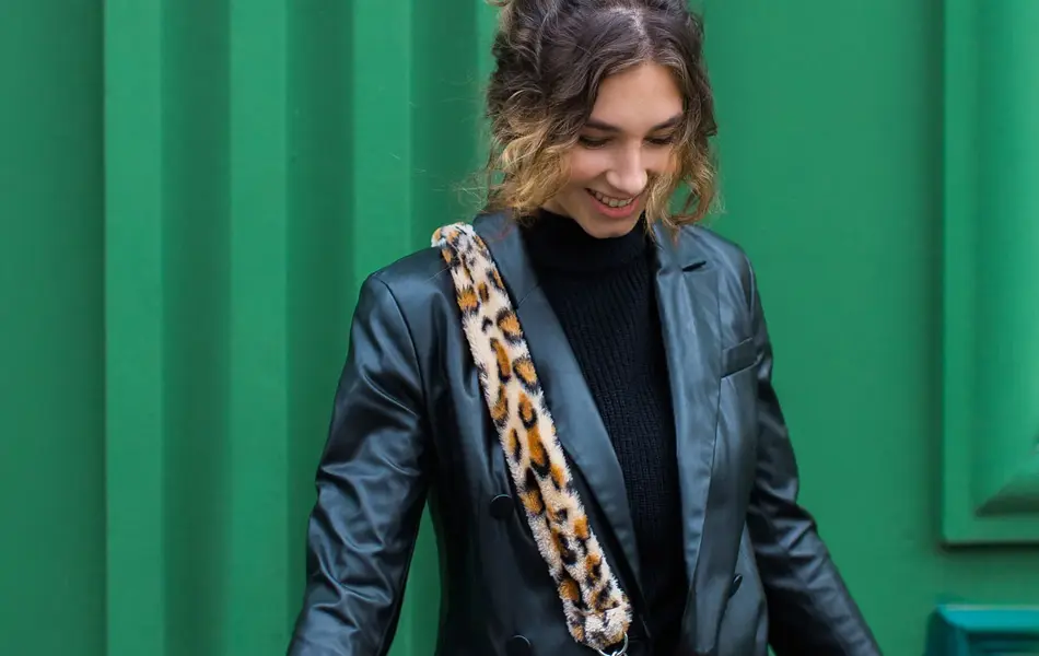 En dame med svart skinnjakke og fluffy veske i leopardmønster står foran en grønn vegg