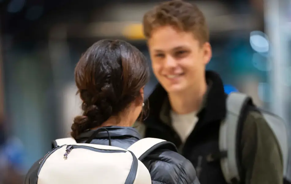 Tenåringer, jente med ryggen mot kamera med boblejakke og lyse beige skolesekk, ser på gutt med jakke og sekk.