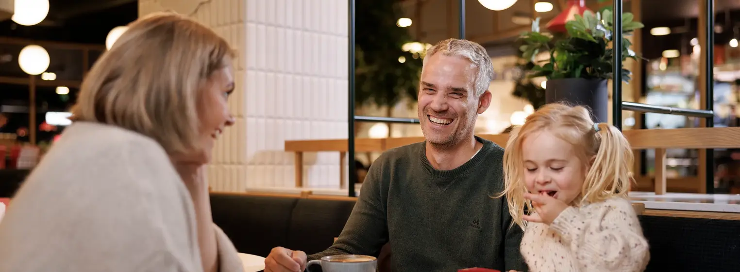 En mann, en kvinne og et barn sitter ved et bord på en kafé, mannen og kvinnen smiler til hverandre