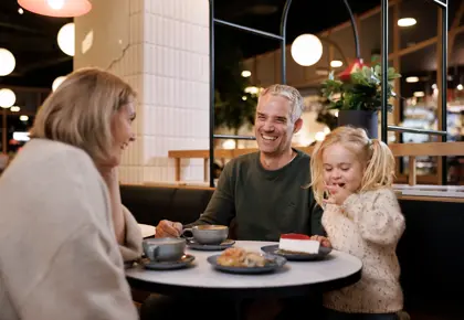 En mann, en kvinne og et barn sitter ved et bord på en kafé, mannen og kvinnen smiler til hverandre