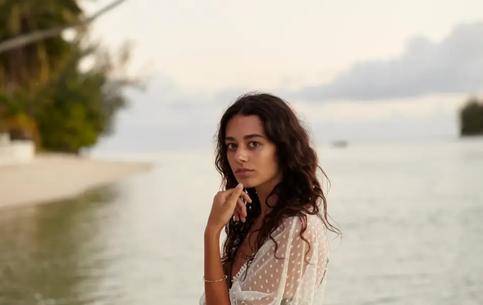 Young brunette stands in the Tahitian ocean at dusk and puts her hand to her chin in an attractive pose wearing cream colored swimwear.