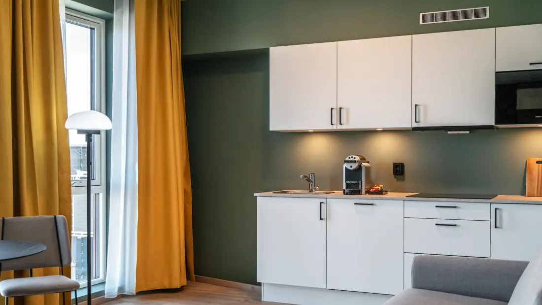 Business rom med kjøkken krok, stort vindu, gule gardiner, grønne vegger og parkett.