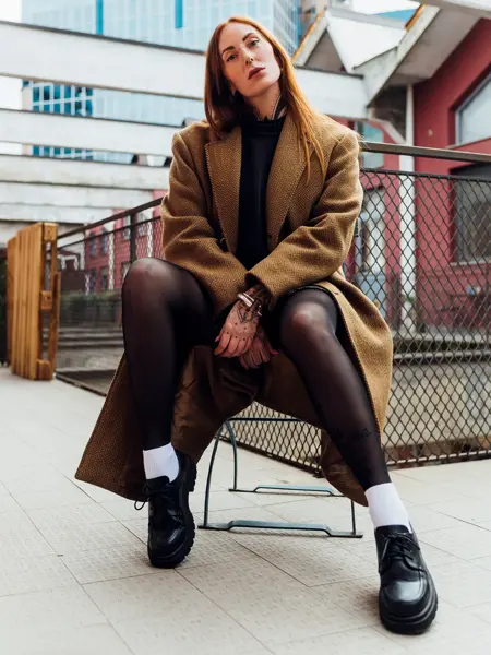 Dame med brun kåpe, sort strømpebukse og sorte snøresko sitter på stol utendørs og poserer for kamera 