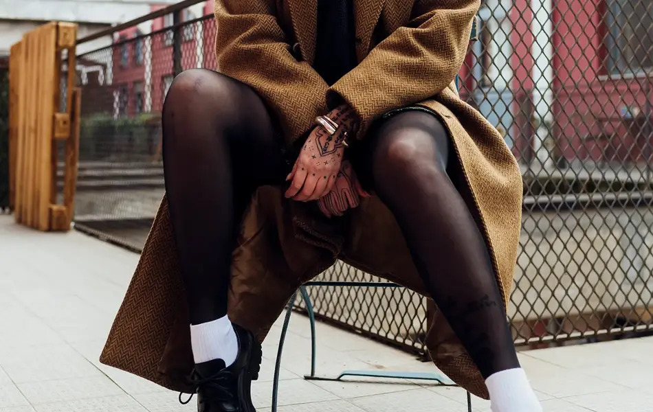 Dame med brun kåpe, sort strømpebukse og sorte snøresko sitter på stol utendørs og poserer for kamera 