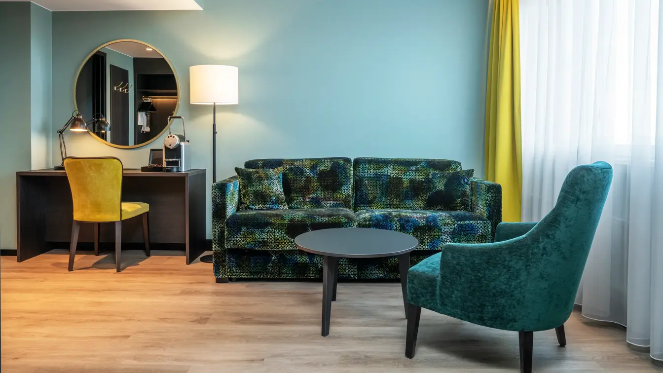 Stue, sofa, salongbord, grønn lenestol, gule gardiner, vindu, TV, parkettgulv, skrivebord, speil på veggen, gul skrivebordstol, gulvlampe 