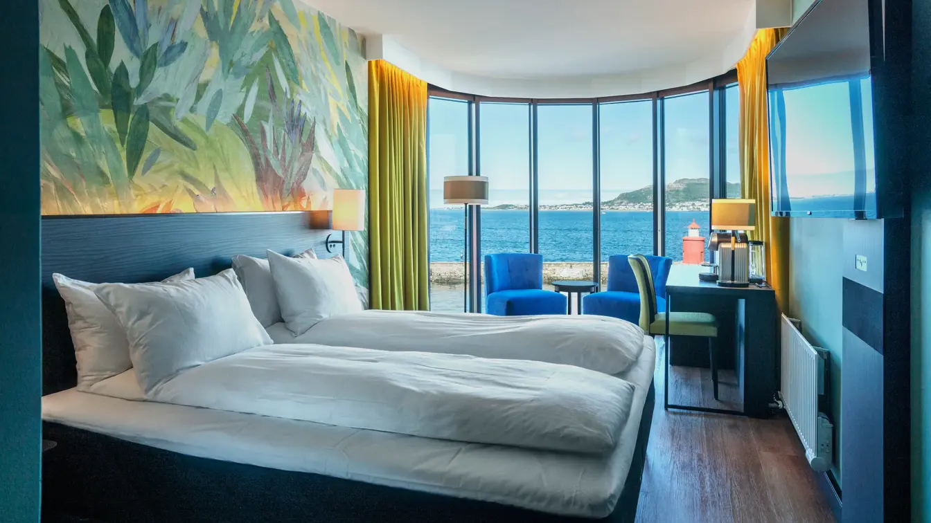 Buevindu med utsikt over vannet og blå himmel, tapetvegg, TV, dobbeltseng, skrivebord, to blå lenestoler