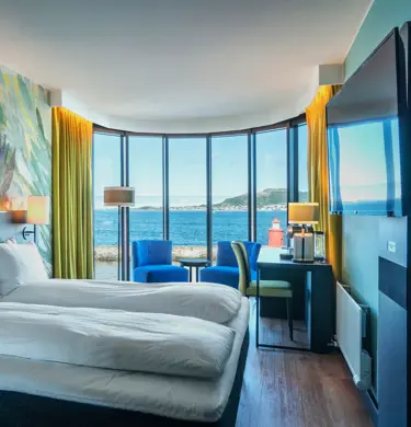 Buevindu med utsikt over vannet og blå himmel, tapetvegg, TV, dobbeltseng, skrivebord, to blå lenestoler