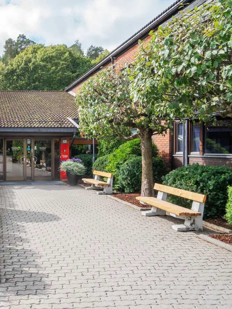 Langt inngangsparti med steinheller, trebenk på høyre side, busker på venstre side, to glassskyvedører i enden av inngangen.