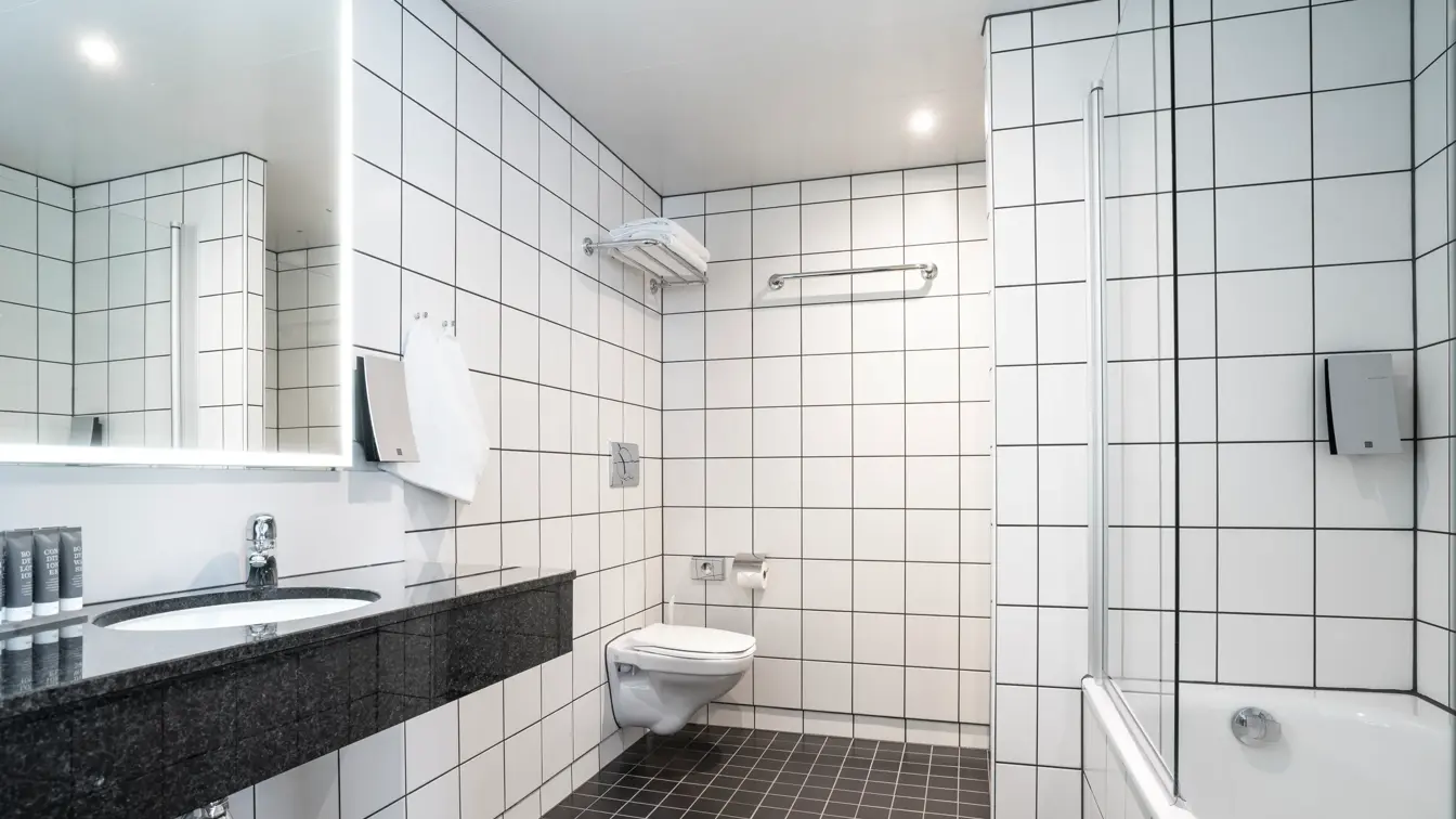 Servant i marmorbenk, speil med led lys rundt, vegghent toalett, badekar, hvite fliser på veggene og mørke på gulvet på bad i suite på Thon Hotel Kirkenes