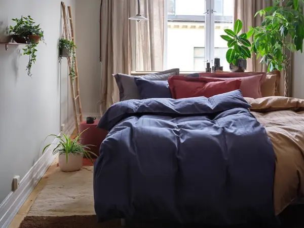 Soverom med oppredd dobbeltseng, teppe på gulvet, vindu med gardiner og grønne planter