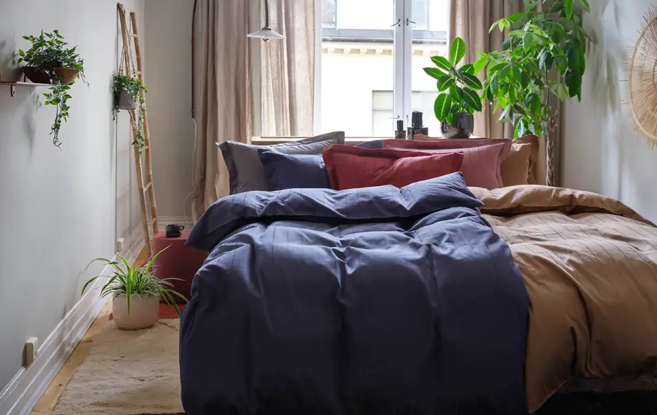 Soverom med oppredd dobbeltseng, teppe på gulvet, vindu med gardiner og grønne planter