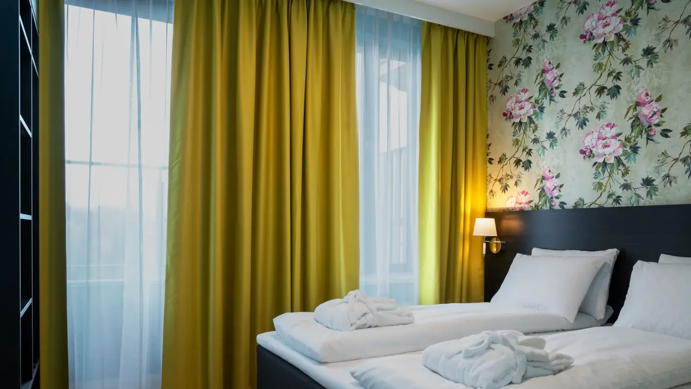 Dobbeltseng, gule gardiner, vindu fra gulv til tak, blomstrete tapetvegg.