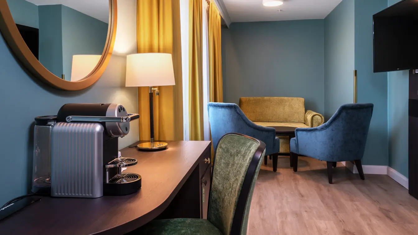 Skrivebord, grønn stol, kaffemaskin, rundt veggspeil, TV, gul sofa, 2 blå lenestoler, vindu, gule gardiner, grønn vegg