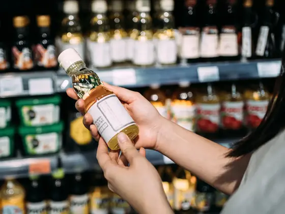 Beskåret bilde av ung asiatisk kvinne som handler dagligvarer i et supermarked. Står ved en frukt- og grønnsaksavdeling, holder en flaske med økologisk matolje og leser næringsinnholdet på etiketten.