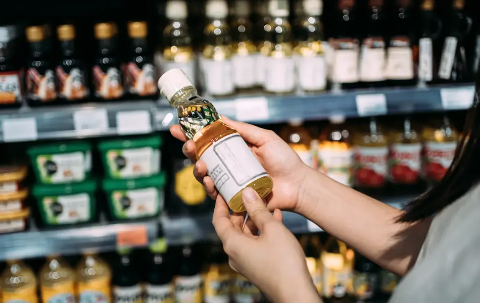 Beskåret bilde av ung asiatisk kvinne som handler dagligvarer i et supermarked. Står ved en frukt- og grønnsaksavdeling, holder en flaske med økologisk matolje og leser næringsinnholdet på etiketten.