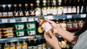 Beskåret bilde av ung asiatisk kvinne som handler dagligvarer i et supermarked. Står ved en frukt- og grønnsaksavdeling, holder en flaske med økologisk matolje og leser næringsinnholdet på etiketten.