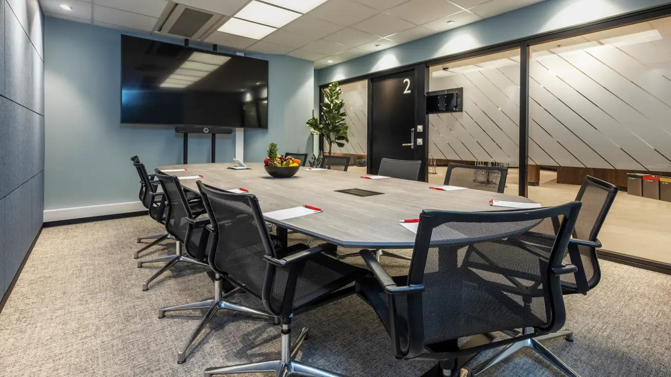 Møterom med styrebords oppsett til 10 personer. Sort TV skjerm, grønn plante i hjørnet og vinduer langs den ene veggen.