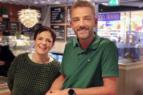 Dame og mann smiler til kamera med Rema 1000 i bakgrunnen